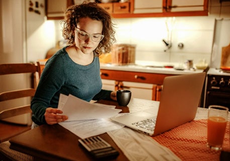 Young woman is sitting at her kitchen table reviewing documents while using a calculator and laptop
